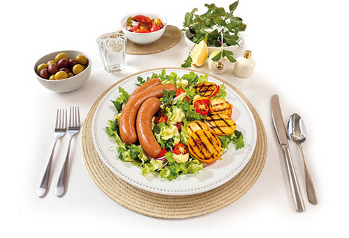Teller mit Würstchen auf Salat, angerichtet mit gegrilltem Gemüse, Besteck, Oliven, Tomaten und Dekoration auf hellem Hintergrund.
